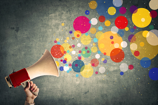 Hand holding a bull horn with cartoon circles of various colors coming out of the horn's bell.