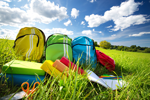 Collection of backpacks, binders, notebooks, and other school supplies laid out in a green field with blue skies above.