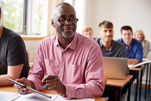 Male of color wearing a pink collared shirt as he sits at a desk in a classroom with others.