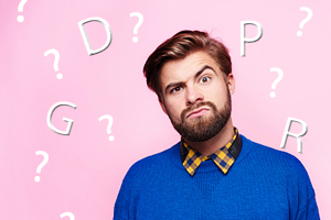 Pink background with the letters, GDPR, and question marks scattered on it and in the foreground is a male in a blue sweater who's looking inquisitively at the camera.