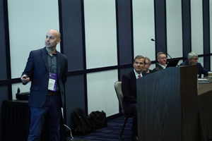 Male speaker standing as he clicks through a presentation while a panel of other individuals are seated next to him.