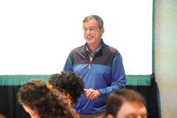 Photo of a smiling male speaker wearing a blue vest.