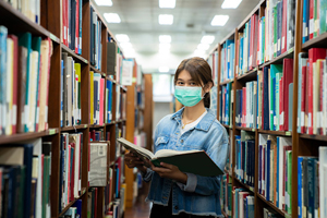Masked student in library holding a book.