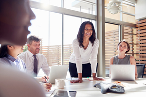 business female smiles and stands over a table with other business people sitting around the table