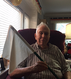 Photo of John Bear holding a white flag while sitting in a red sofa chair. 