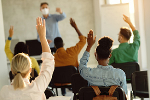 Teacher wearing a mask while standing at the front of a classroom chooses a student from the class who all have their hands raised.