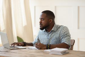 male working on his laptop while sitting at a wooden table in a plain neutral colored room 