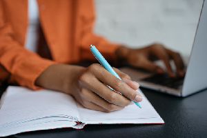 person wearing an orange jacket, taking notes with a blue pen while on their laptop