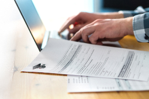 person working on their laptop while at a wooden desk with scattered papers off to the side