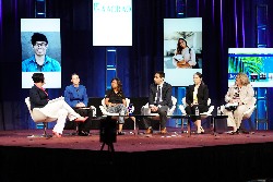 Panel of six people sitting on a stage with multiple screens showing different images behind them.