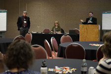 Photo from the audience's perspective of three figures speaking from the front of an event space.