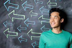Male in a green shirt standing in front of a blackboard with arrows drawn on it all pointing off in the same direction.