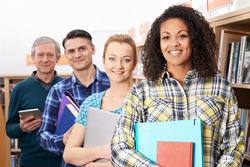 Four people in a line smiling at the camera while holding books and binders in a library.