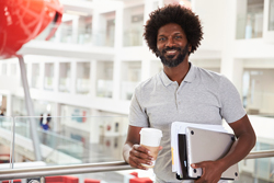 Male of color holding a coffee and folders while smiling at something out of frame.