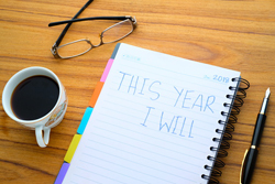 Wooden desk with a coffee mug, glasses, pen, and a notepad with the words "this year I will" written in it all on top of the desk.