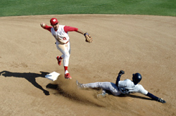 One baseball player in a blue helmet slides toward a base while another player in a red hat throws the ball to someone out of frame.