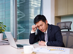 Male sitting at an office desk while he makes an expression of stress and fatigue. 