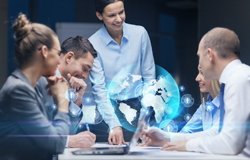 Group of business professionals talking amongst themselves with a hologram of the globe visible in the center of them.