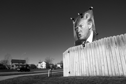 Black and white photo of a corrugated steel wall with a large photo of Donald Trump perched on top.