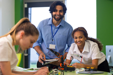 Smiling teacher helps a student with what appears to be a robot while another students works diligently in the foreground.