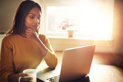 Female of color using her laptop while light pours in through her window.