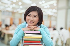 Female of asian origin smiling as she leans on a stack of books.