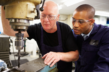 A student looks on as an instructor demonstrates how to use a drill press.