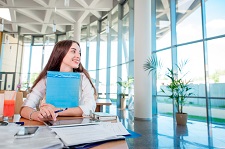 Female student smiling as she studies in a room with floor to ceiling windows. 