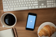 Photo of a wooden surface with a mug of coffee, smartphone, keyboard, and croissant visible. 