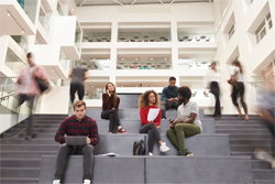 Students sitting and working on the steps of a white atrium with blurred people walking past.