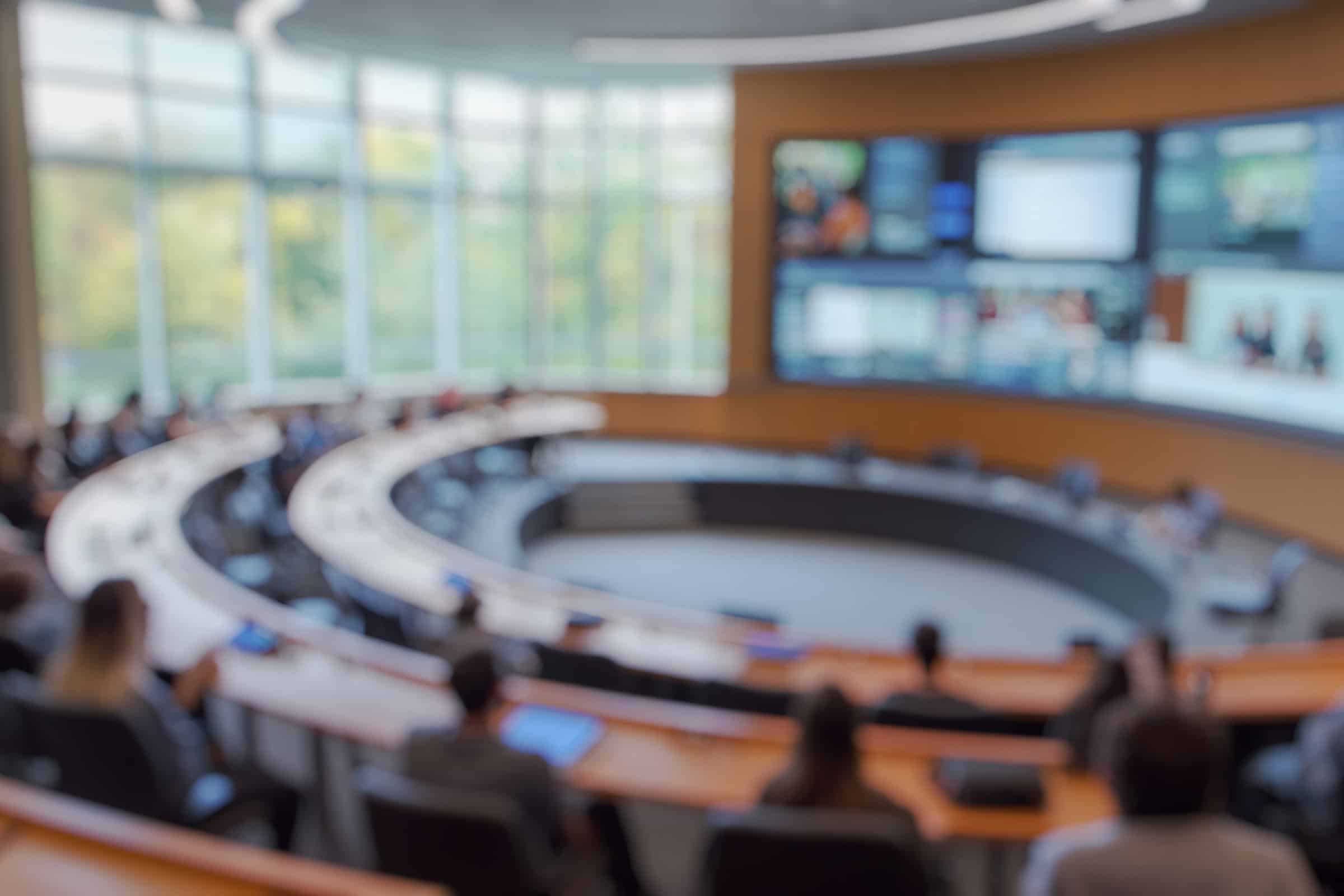 Blurred view of a conference room with people seated and a screen in the background