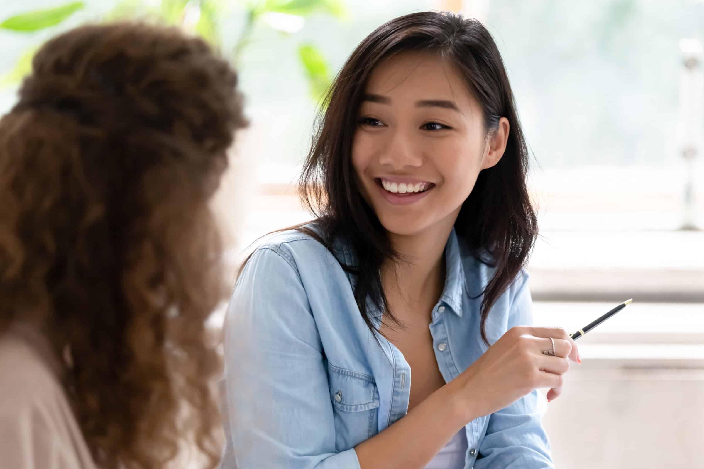 Smiling woman writing with a pen while talking with another person in a bright room