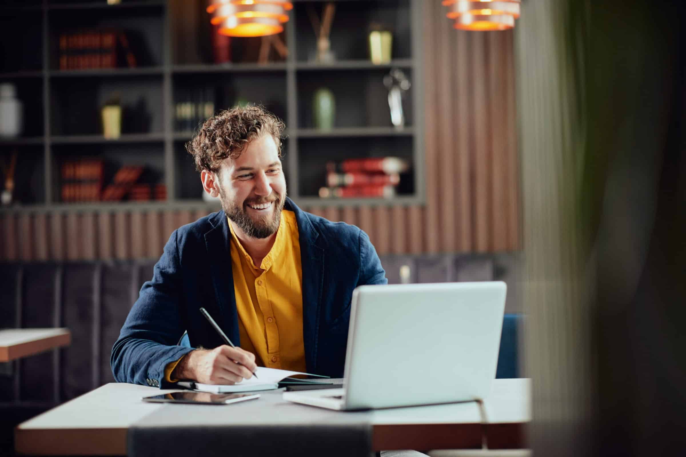 Smiling man working at a desk, writing notes while using a laptop in an office