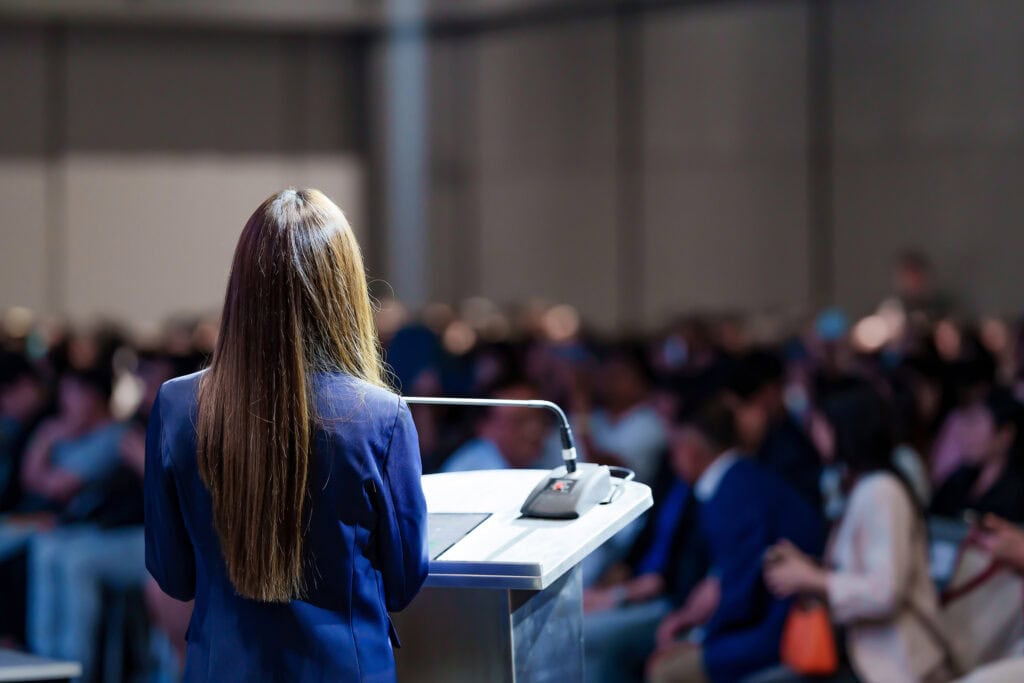Woman in a blue blazer speaking at a podium to an audience in a conference hall