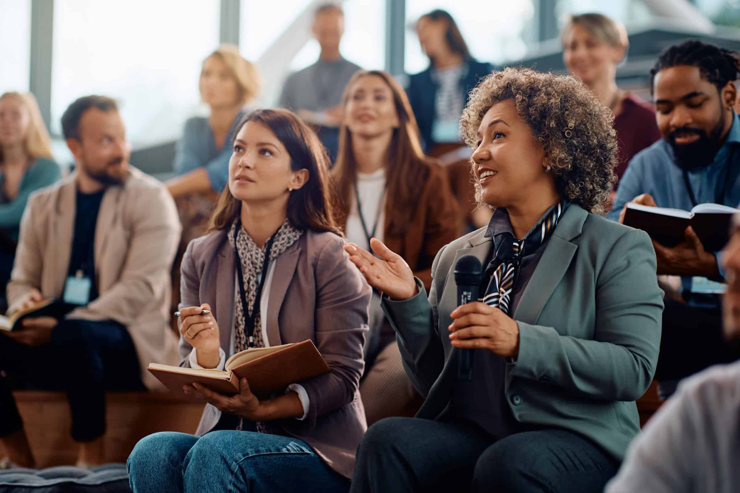 Woman in a suit speaks into a microphone while seated at a panel discussion with other attendees listening