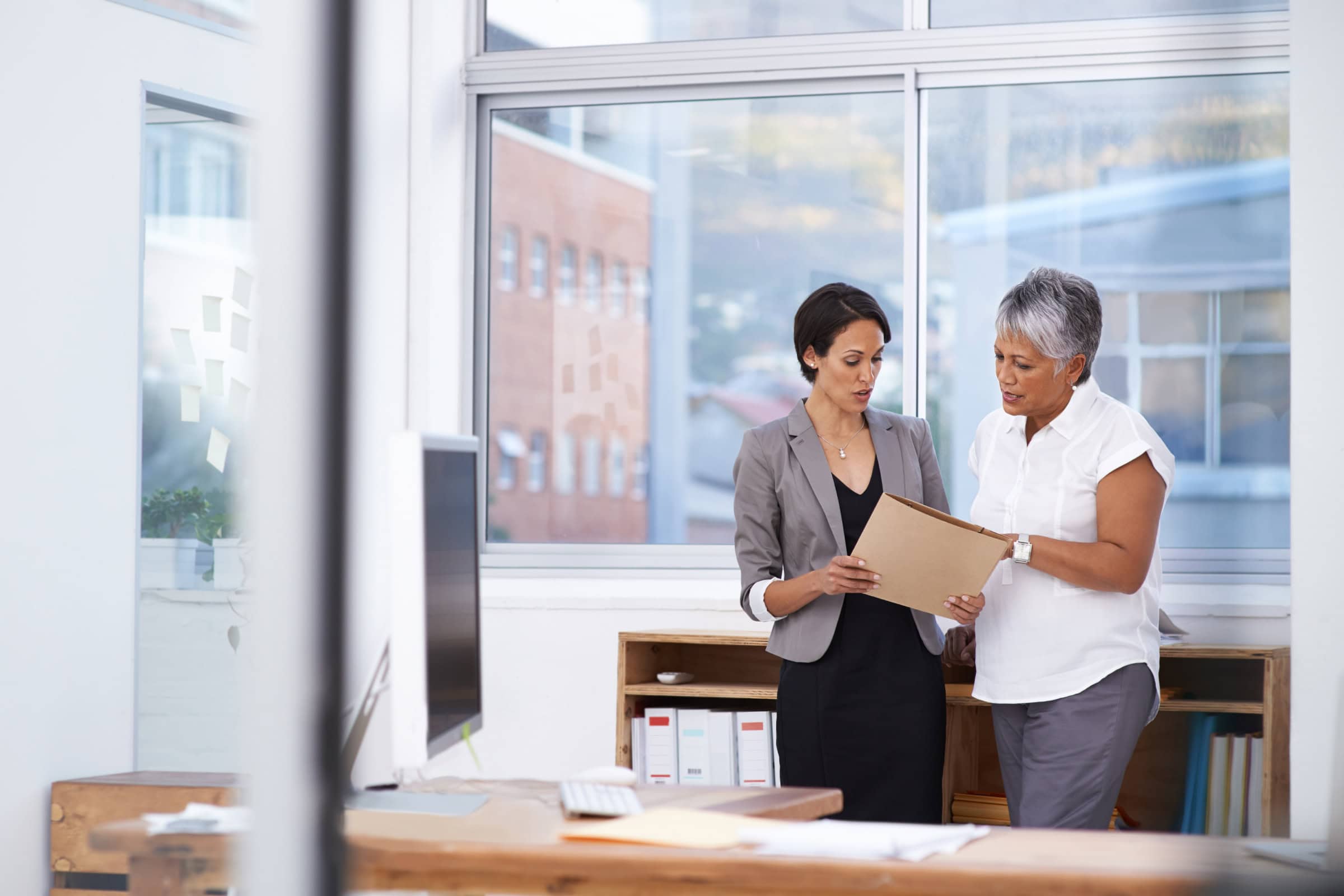 Two office workers reviewing documents while standing near a window in a modern workspace