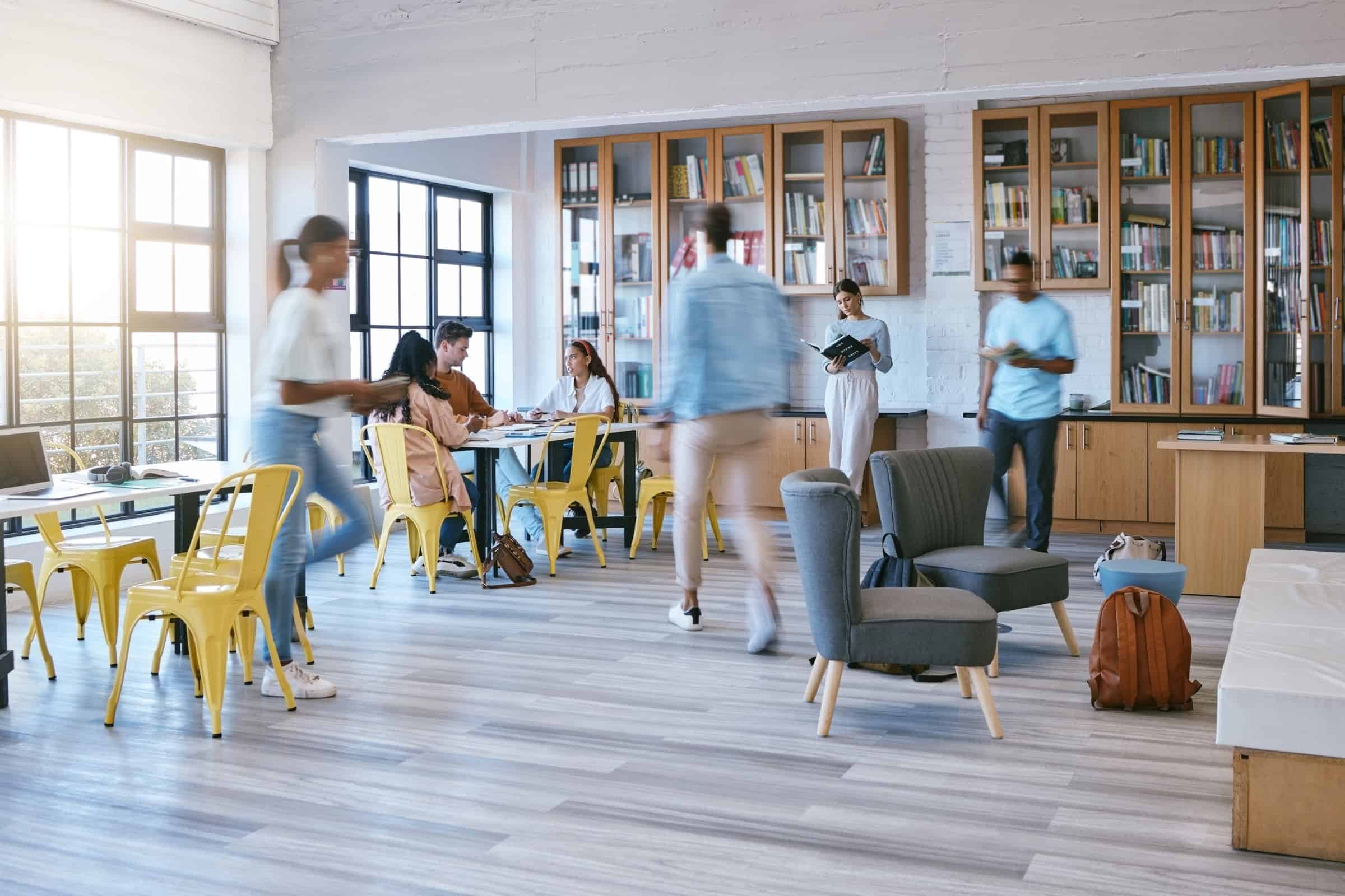 View of a busy room with yellow chairs and tables, books and people moving