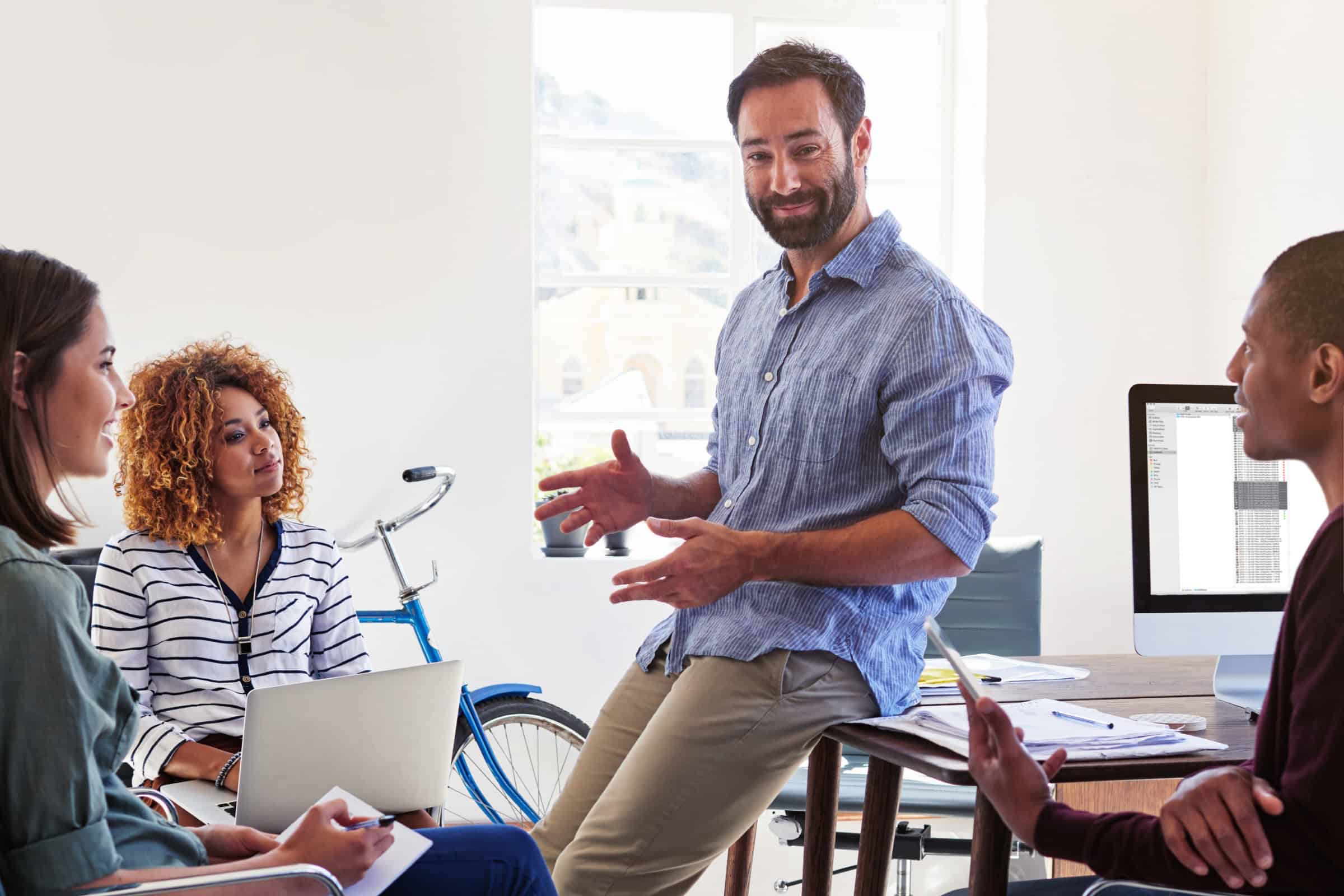Team meeting in an office with a man presenting while colleagues listen and work at laptops