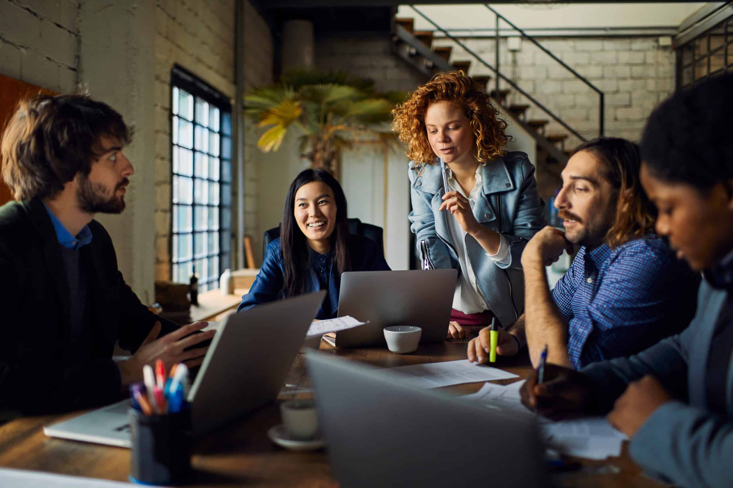A team of coworkers discussing ideas around a table with laptops in a meeting room