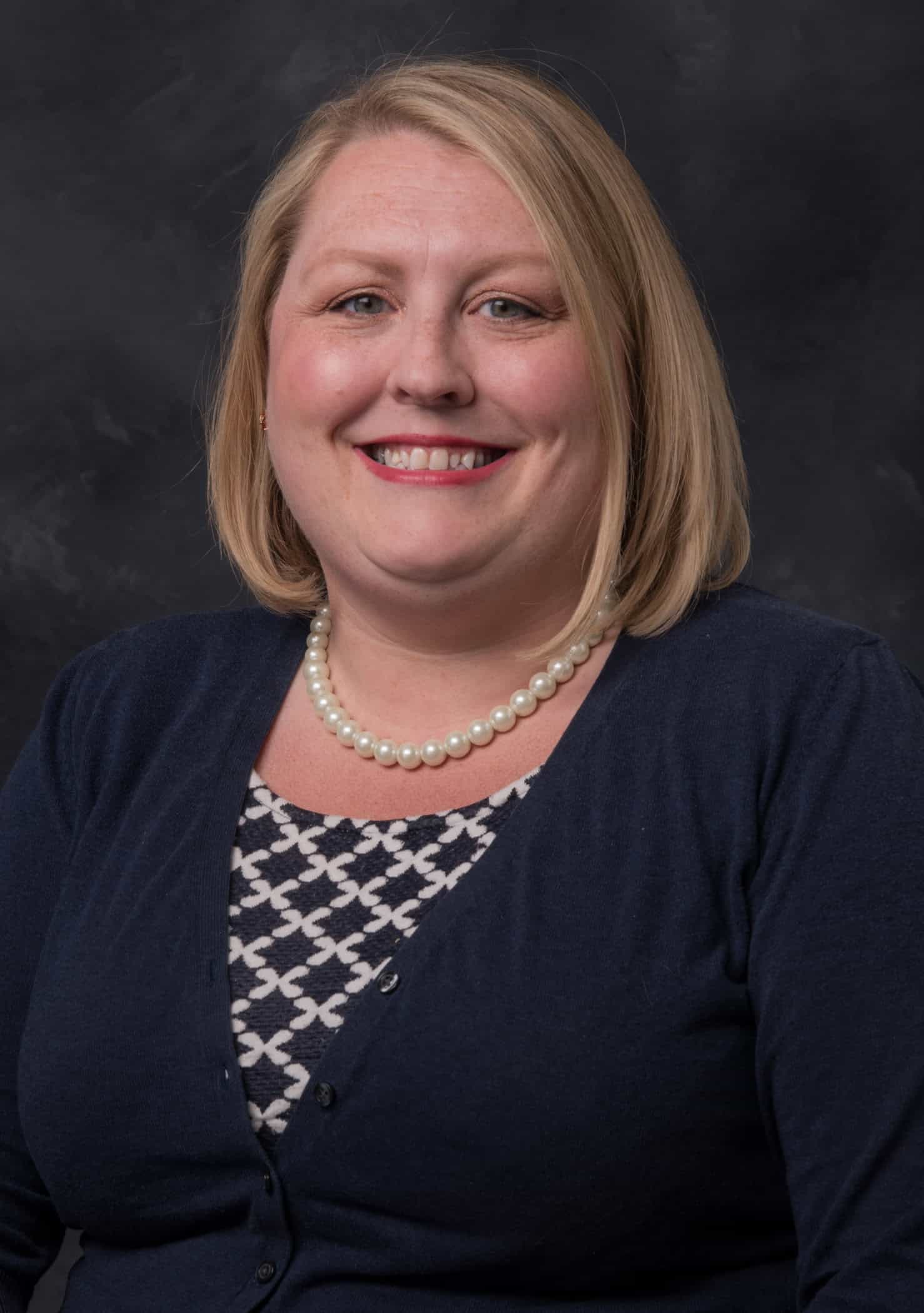 Portrait of a smiling woman wearing a pearl necklace and navy cardigan against a dark background