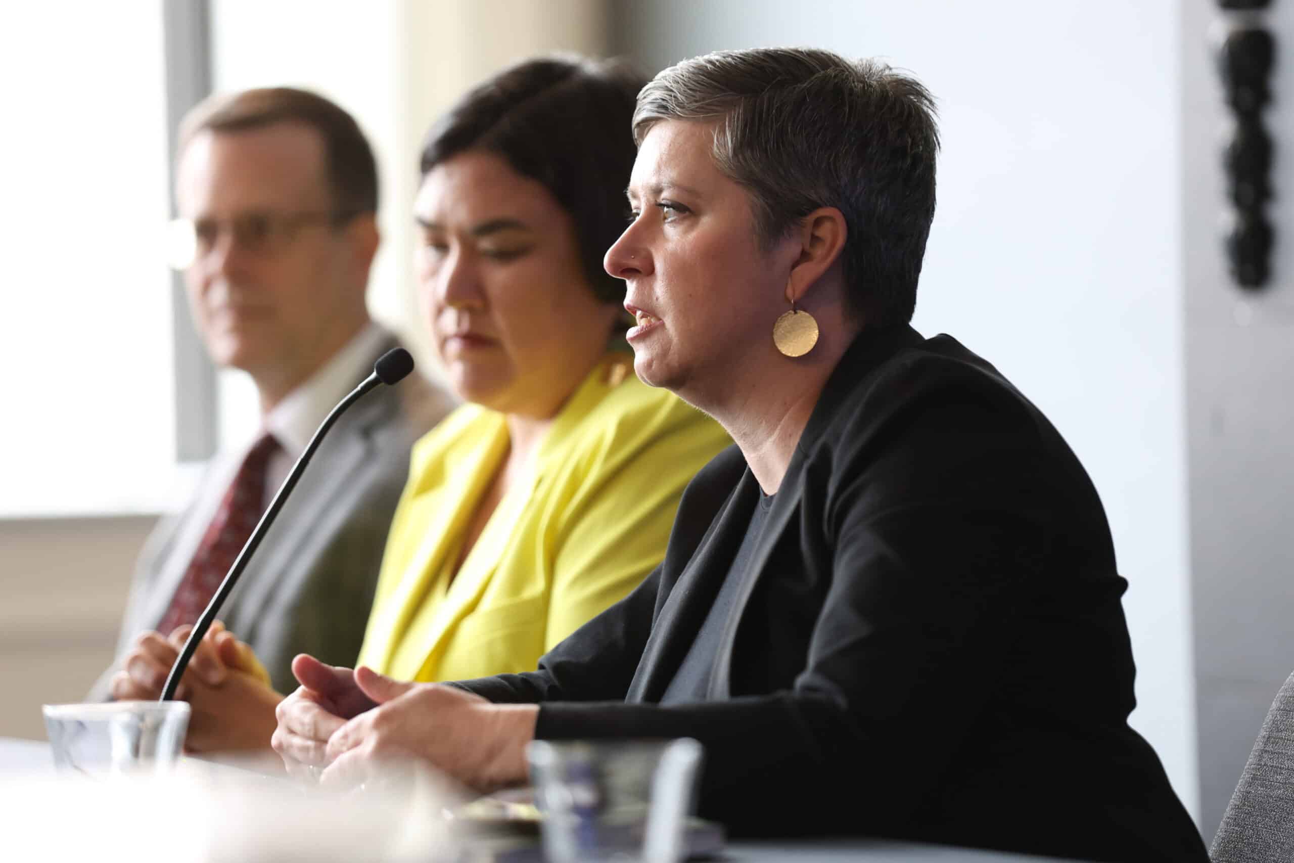 Three panelists seated at a conference table with a microphone. A woman in a black blazer speaks; a woman in yellow and a man in a suit sit behind her