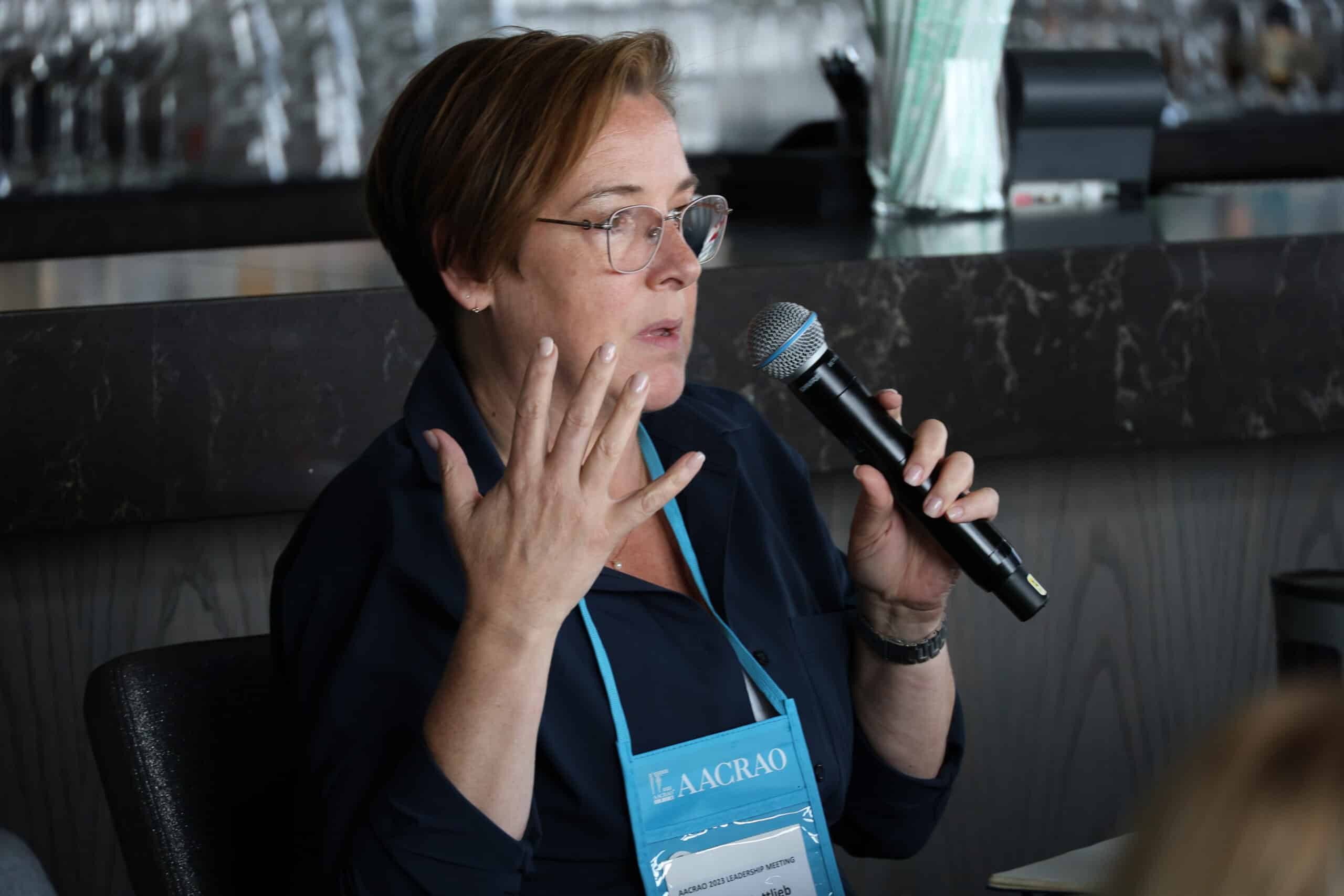 woman with glasses and short hair holds a microphone and speaks