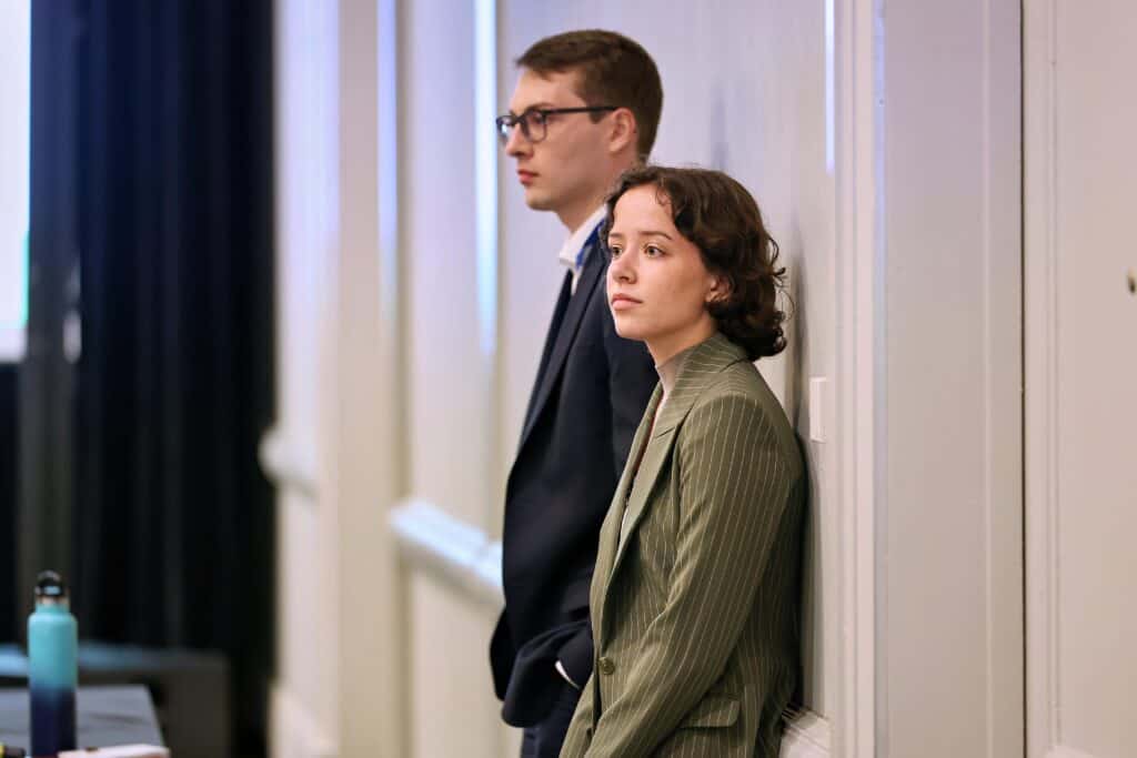 Two people in business attire standing against a wall in a hallway, looking ahead