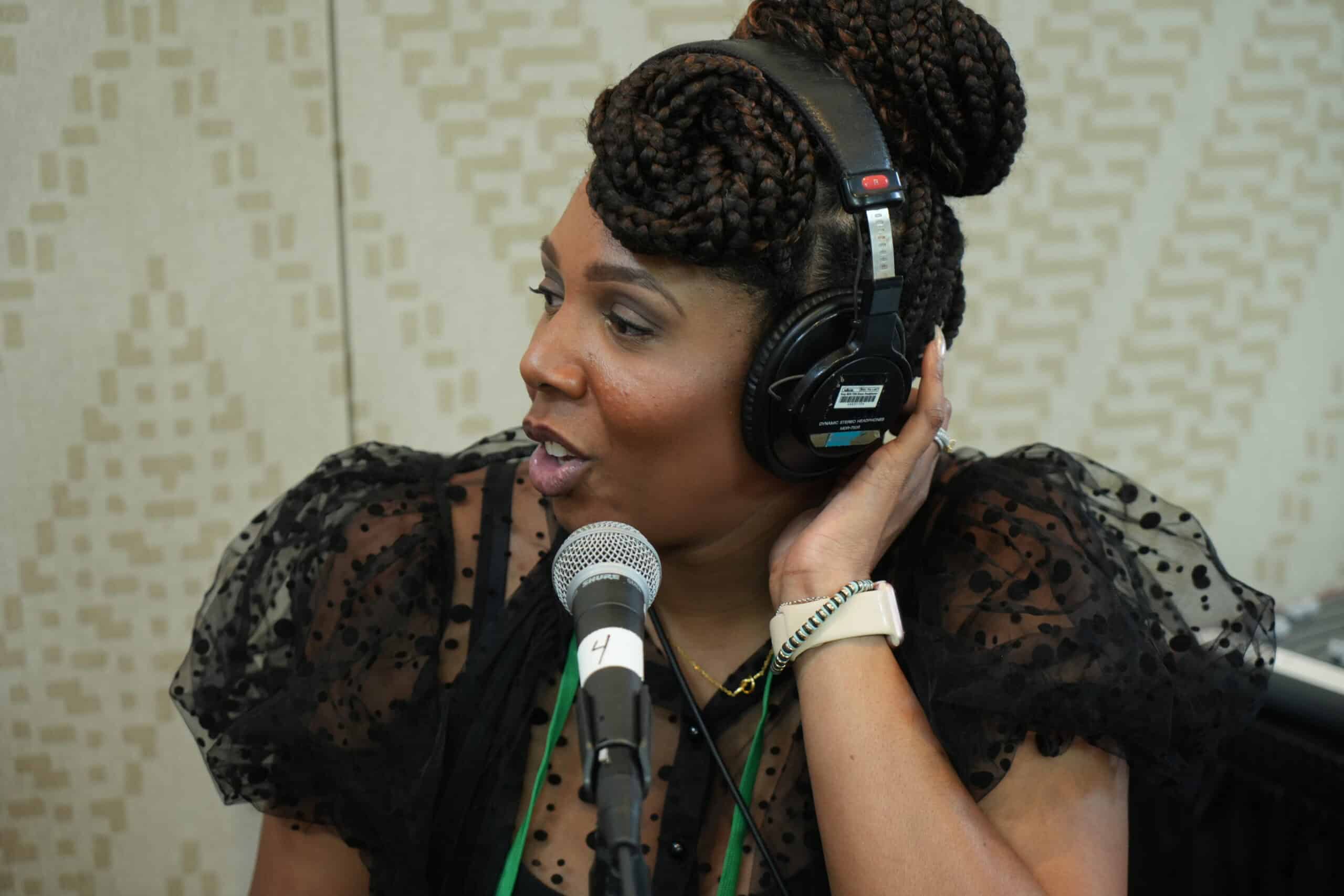 Woman with dark sking and hair up wears headphones and speaks to a microphone while looking to the left
