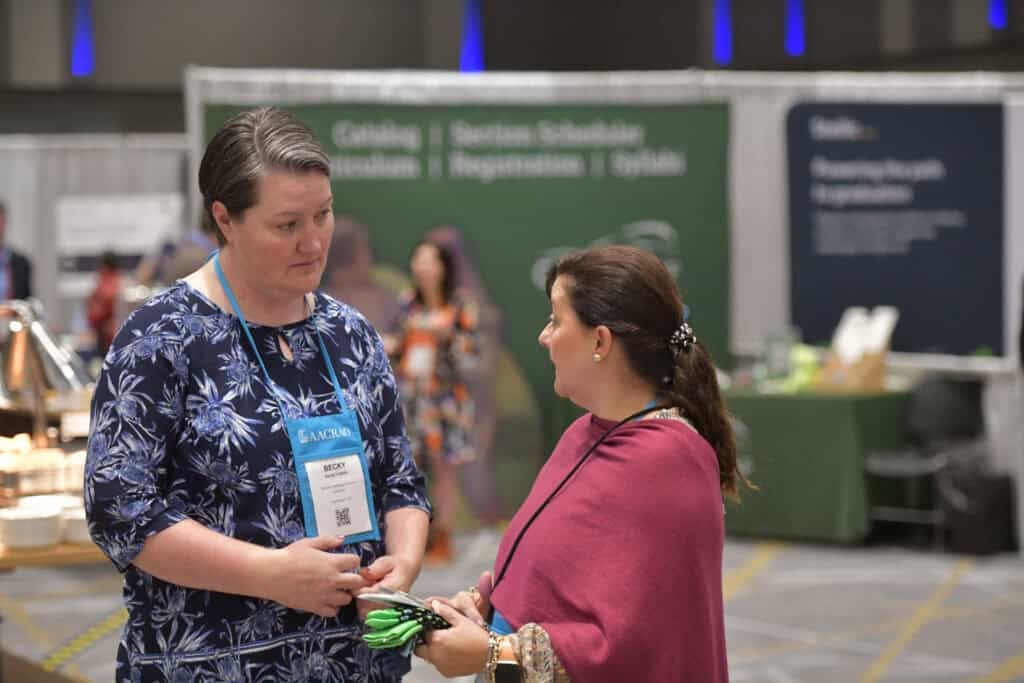 Two conference attendees wearing AACRAO lanyards speak with each other on a convention floor. A green registration signage backdrop is visible behind them.