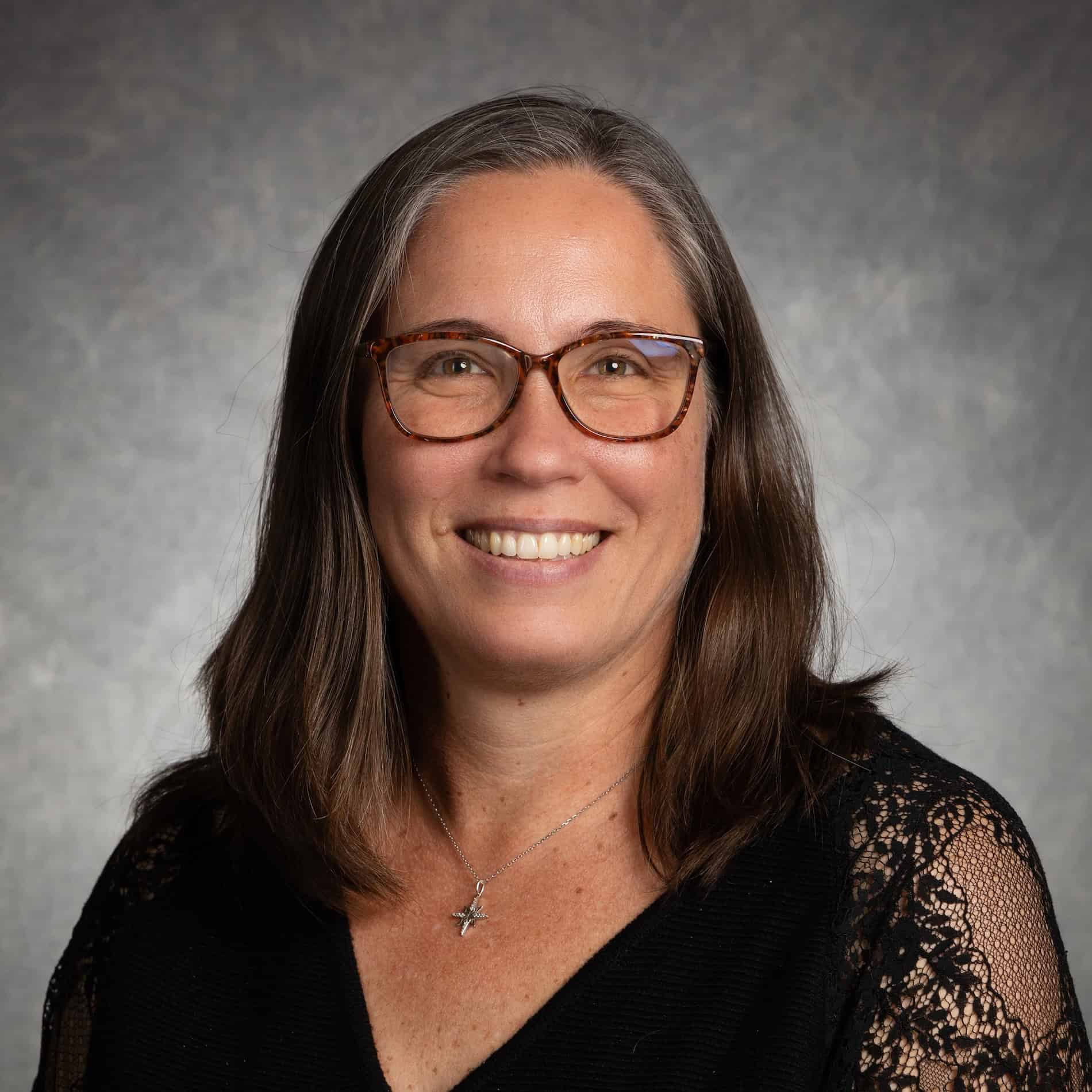 Portrait of a smiling woman wearing glasses and a black lace top against a gray background