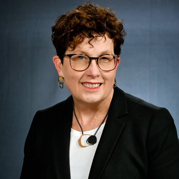 Portrait of a smiling woman wearing glasses and a black blazer against a blue-gray background