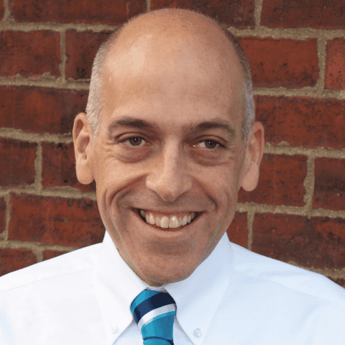 Portrait of a smiling bald man in a white shirt and blue striped tie against a brick wall