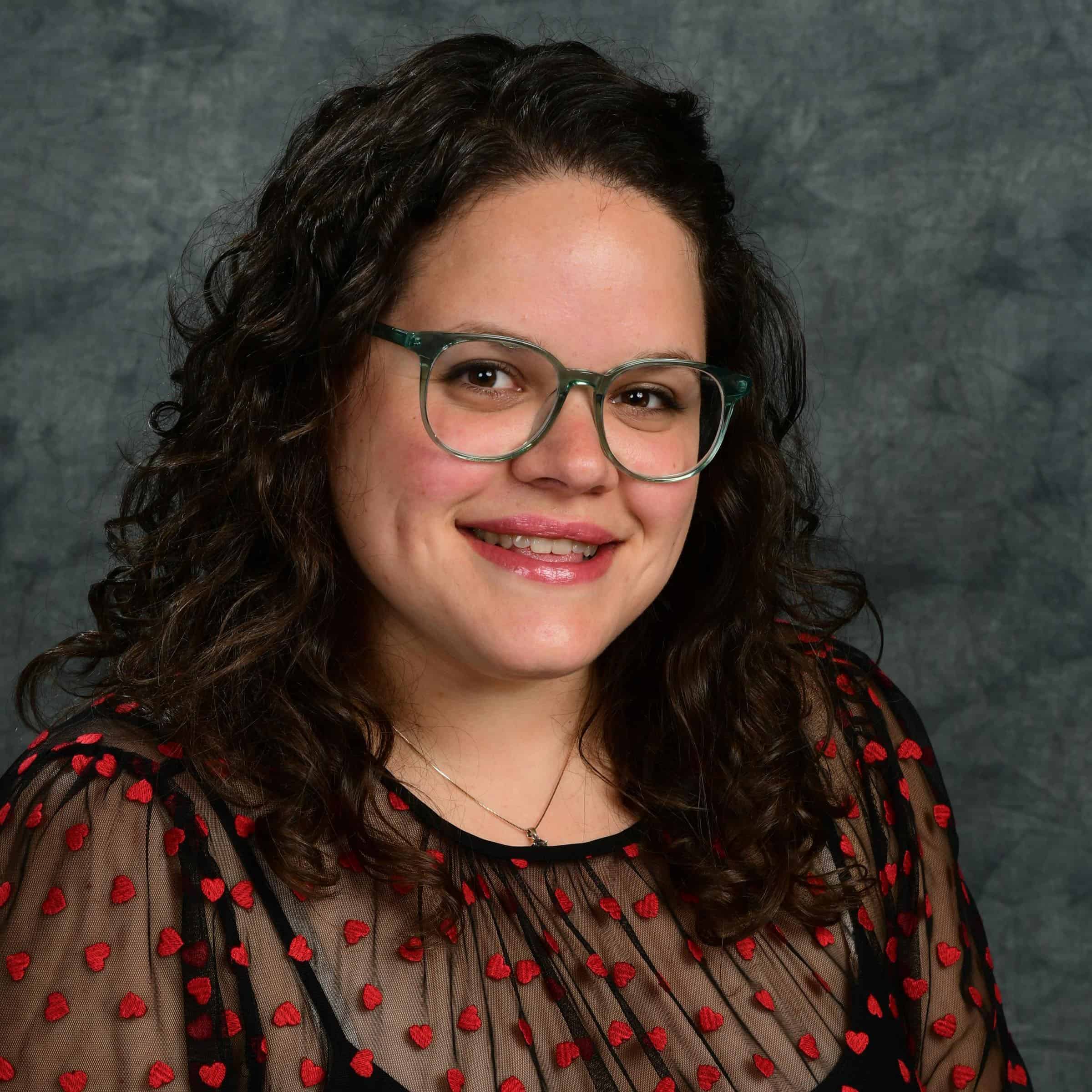 Portrait of a smiling woman with curly hair and glasses against a gray backdrop