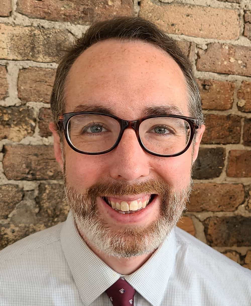 Smiling man with glasses and a beard in a dress shirt and tie against a brick wall